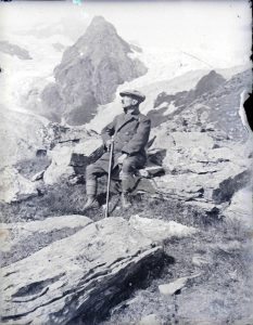 Un signore in tenuta da montagna si gode, seduto su una roccia, lo spettacolo delle montagne innevate negli Anni '20