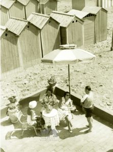 Tre signore sedute al tavolino di un bar sulla spiaggia si intrattengono con un signore in costume da bagno. Sullo sfondo le cabine. Siamo negli Anni '50