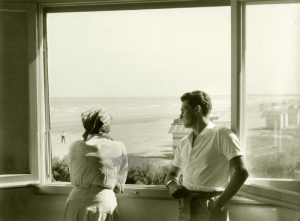 Una giovane coppia affacciata ad una finestra guarda il mare e la spiaggia ancora fuori stagione. Anni '50