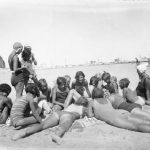 Un gruppo di giovani bagnanti con abbigliamento marino proprio degli Anni '30, distese a chiacchierare sulla spiaggia di Cesenatico. In lontananza il canale con i suoi capanni e le sue vele.