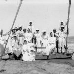 Un nutrito gruppo di bagnanti a Cesenatico, nel 1916, tutti rigorosamente vestiti, seppur in bianco, sono in posa sulla riva del mare, su un pattino