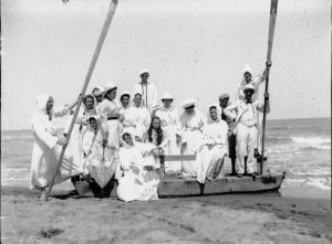 Un nutrito gruppo di bagnanti a Cesenatico, nel 1916, tutti rigorosamente vestiti, seppur in bianco, sono in posa sulla riva del mare, su un pattino